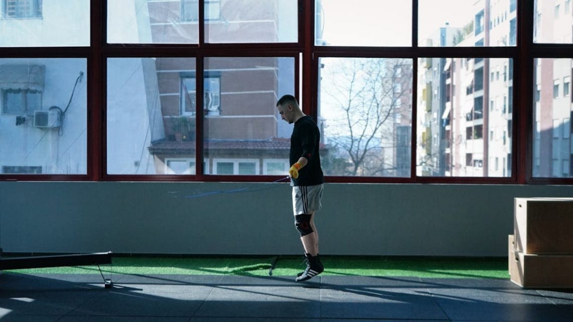 Young man exercising with a skipping rope in an urban gym with large windows and city view.